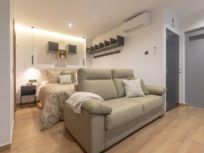 Una sala de estar moderna con un sofá, una cama con ropa de cama estampada, una mesita y un aire acondicionado instalado en la pared.
