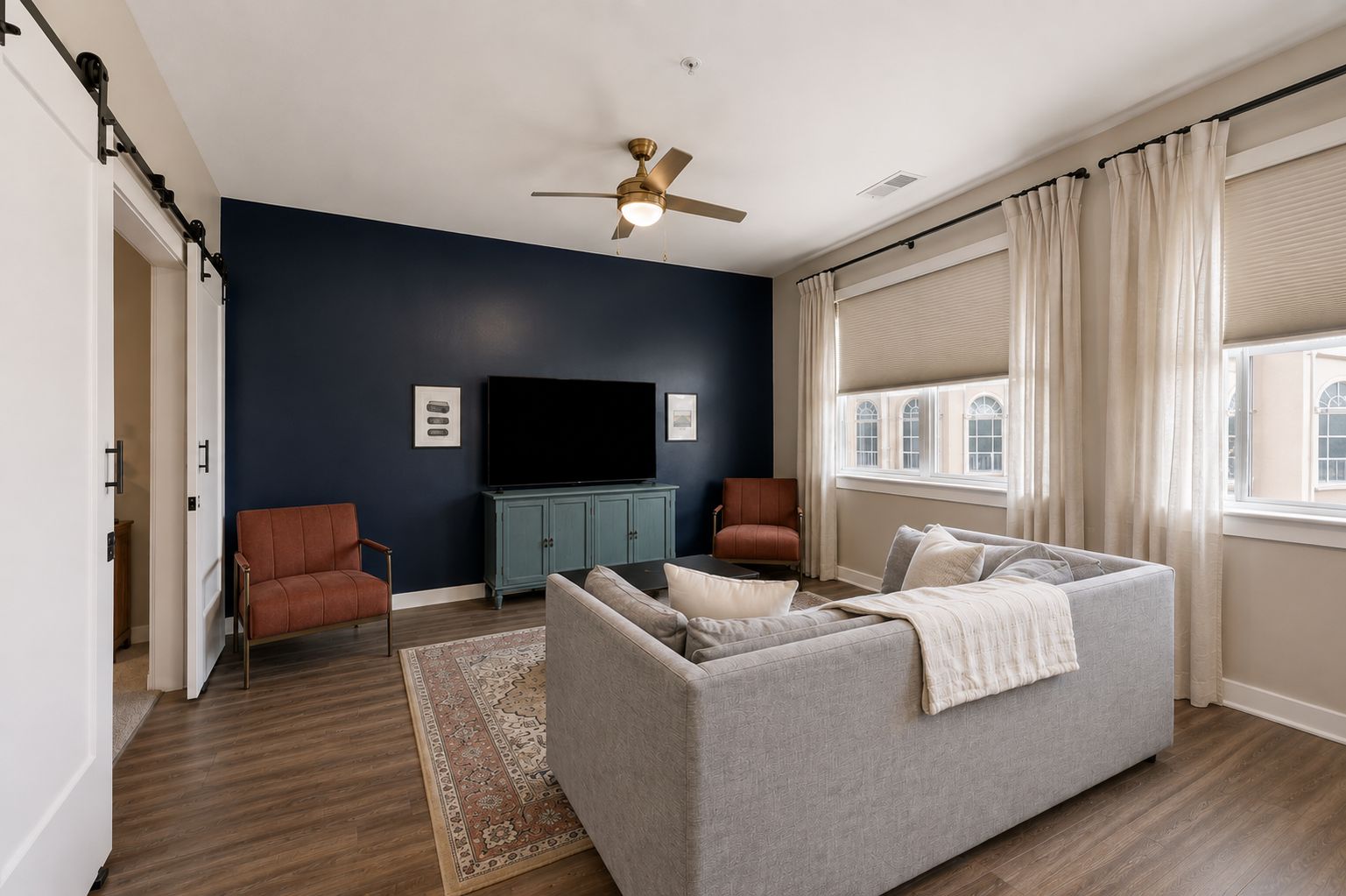 A living room with a grey sofa, two armchairs, a flat-screen TV, a ceiling fan, and a window with curtains.