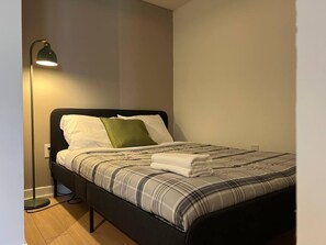 A single bed with a plaid bedspread, a green pillow, and a black headboard. A floor lamp is positioned to the left of the bed. - StayToronto - The CityPlace Escape (Toronto)