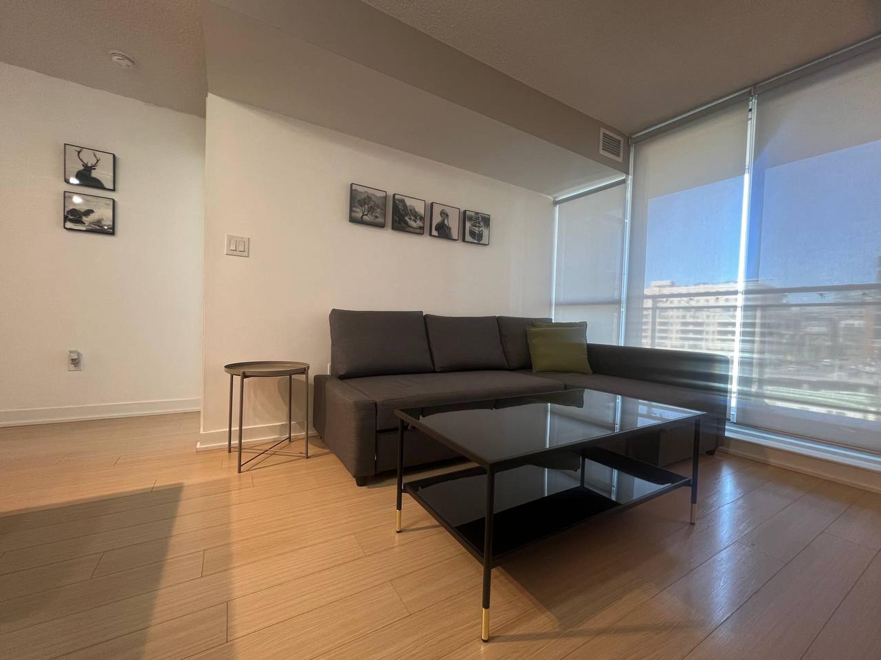 A modern living room with a grey sofa, a glass coffee table, and a small side table. There are framed pictures on the wall and a large window providing a view of the city.