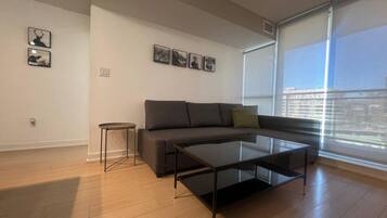 Uma sala de estar moderna com um sofá cinzento, uma mesa de centro de vidro e uma pequena mesinha lateral. Quadros emoldurados adornam a parede, e uma grande janela oferece vista da cidade.
