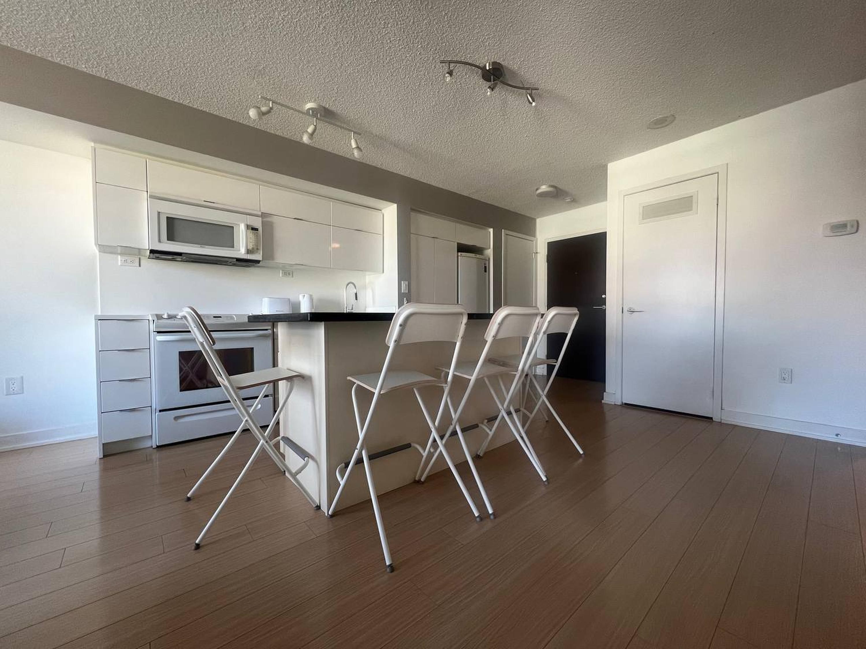 A modern kitchen with white cabinets, a microwave, and a stainless steel oven. There are three white chairs and a small table.
