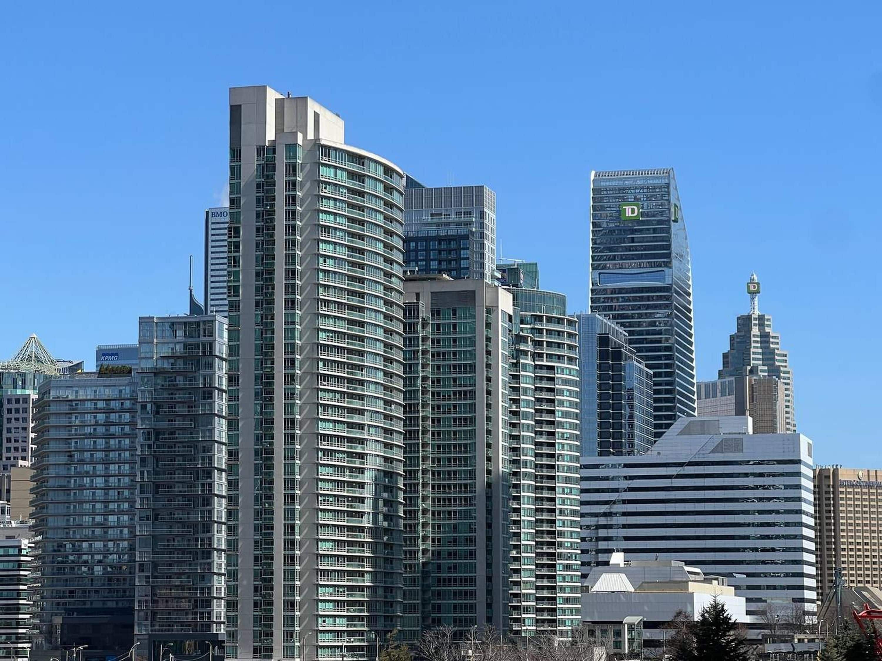 Une vue sur les toits d’une ville, avec des immeubles modernes de grande hauteur, dont un bâtiment à la forme incurvée et un autre, plus élevé, surmonté d’une flèche.