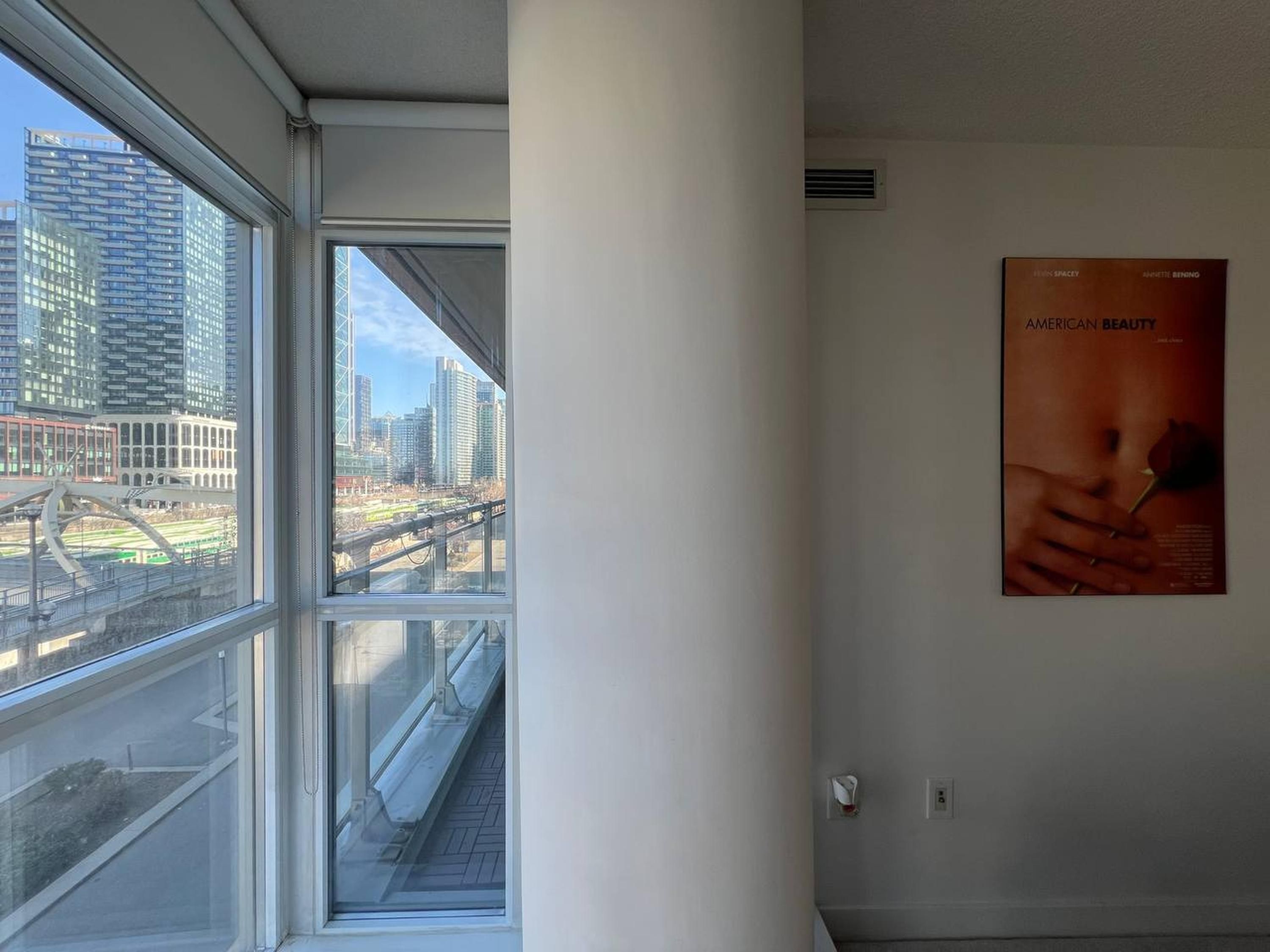 A room with a large window offering a view of a cityscape, a poster on the wall, and a visible fire alarm.