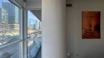 Quarto com uma grande janela que oferece vista para a paisagem urbana, um pôster na parede e alarme de incêndio visível.
