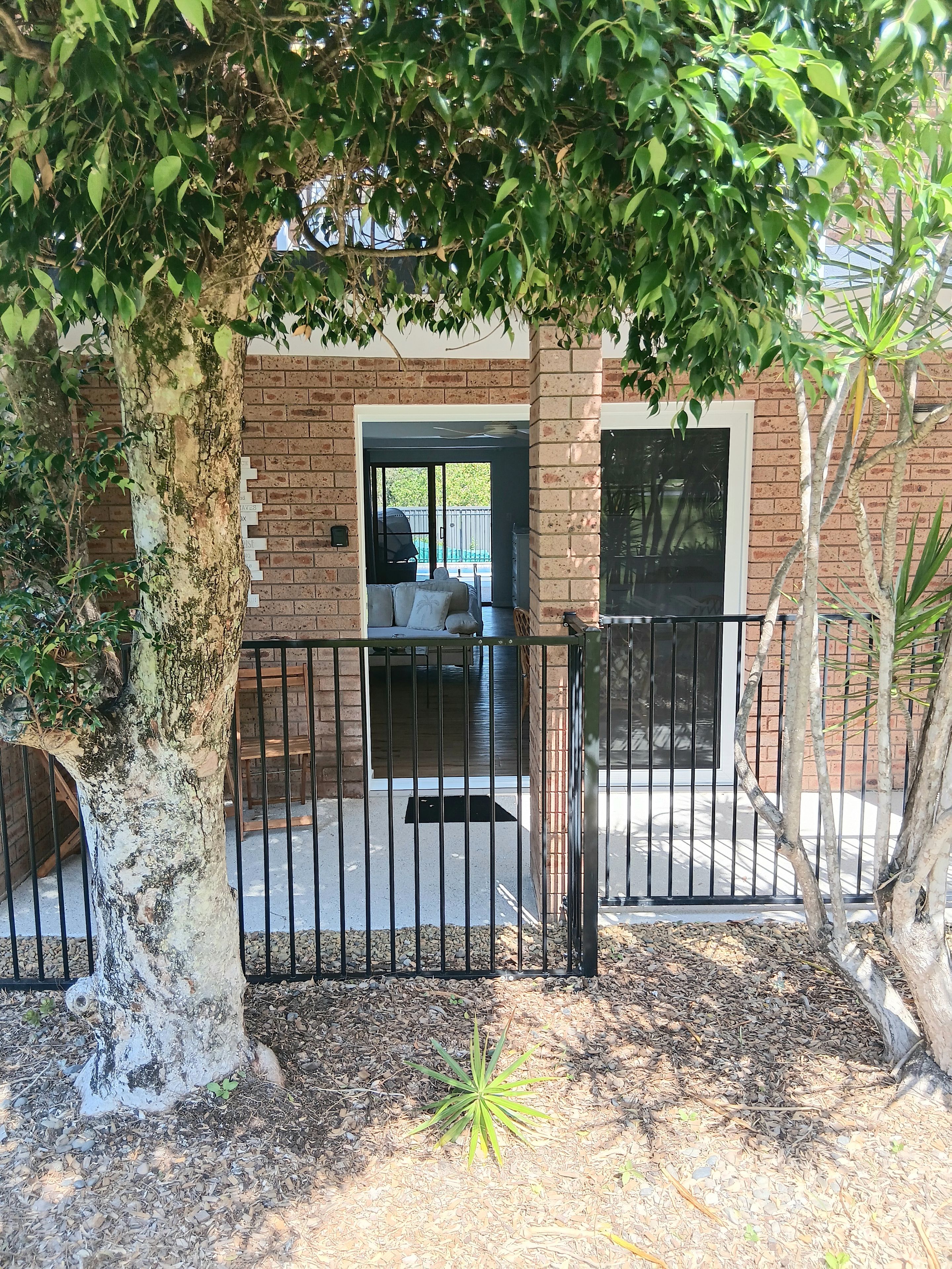 A brick building with a covered patio area, a metal gate, and a tree in the foreground.