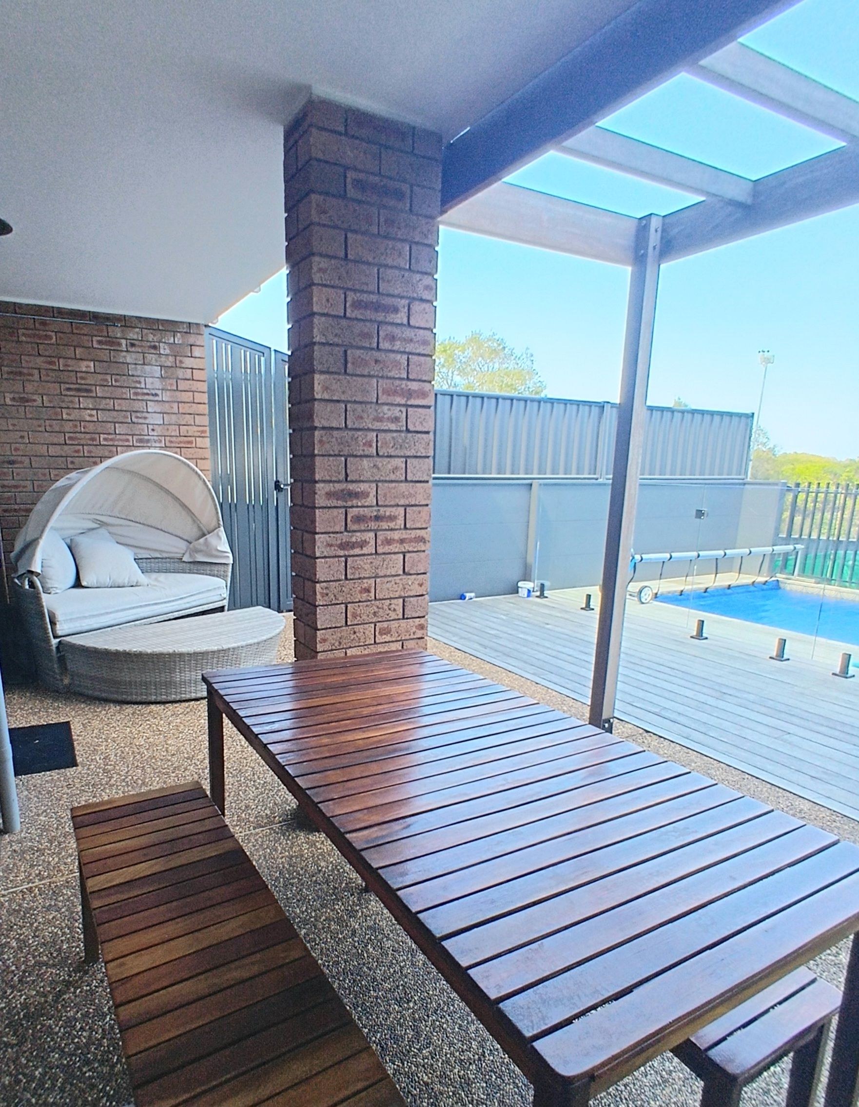 A patio area with a wooden table and bench, a brick pillar, and a view of a swimming pool.