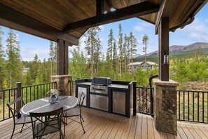 A covered outdoor deck with a grill and seating area, surrounded by trees and a mountain view. - Alpine Exhale: Luxury Mountain Home with amazing views, private hot tub & high end finishes. (Breckenridge)