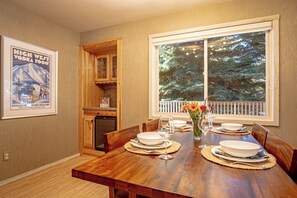 A dining room with a wooden table set for four, a window with a view of trees, and a poster on the wall. - 3BR Elkhorn Village condo with pool, hot tub, fireplace, and mountain access. (Sun Valley)