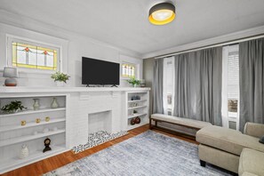 Ein Wohnzimmer mit Kamin, einem flachbildfernseher darüber, einer Couch und einem fenster mit Vorhängen.