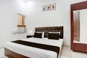 A bedroom with a bed, a nightstand, a wall-mounted air conditioner, a wooden door, and three framed pictures on the wall. - Super Hotel O Mathura-bharatpur (Mathura)