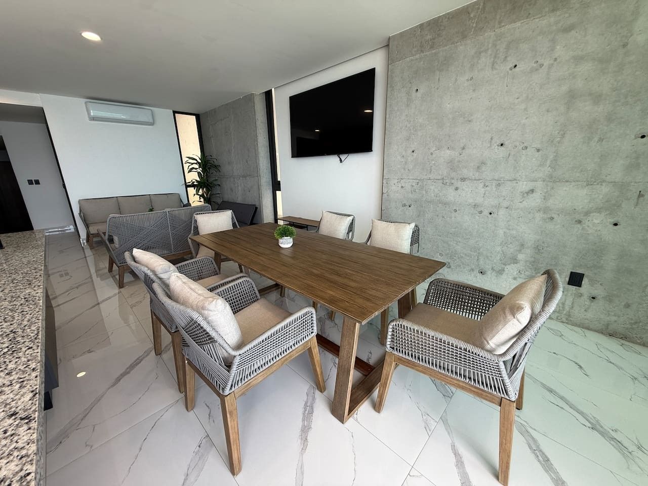 Uma área de jantar moderna com mesa de madeira e cadeiras de vime, televisão de tela plana fixada em uma parede de concreto e um sofá ao fundo.