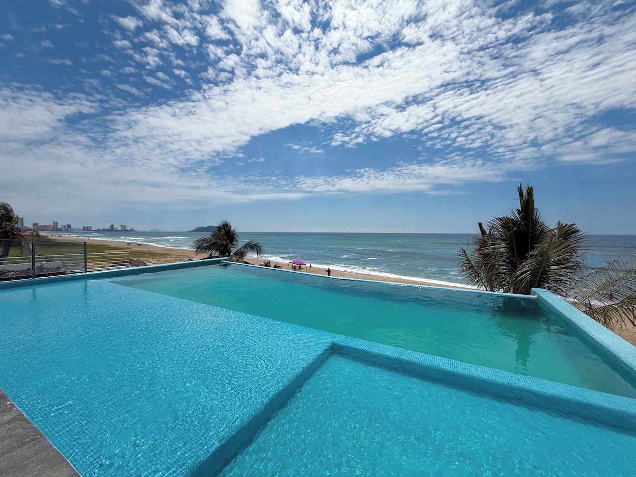 Un cielo azul despejado con nubes, una piscina y una playa al fondo.