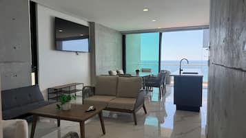 Ein modernes Wohnzimmer mit einer Couch, einem Couchtisch und Sesseln, einem flachbildfernseher an der Wand und Meerblick durch große Fenster.
