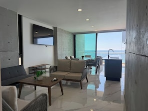 Un salon moderne avec un canapé, une table basse et des fauteuils, un téléviseur à écran plat fixé au mur, et une vue sur l’océan grâce à de grandes fenêtres.