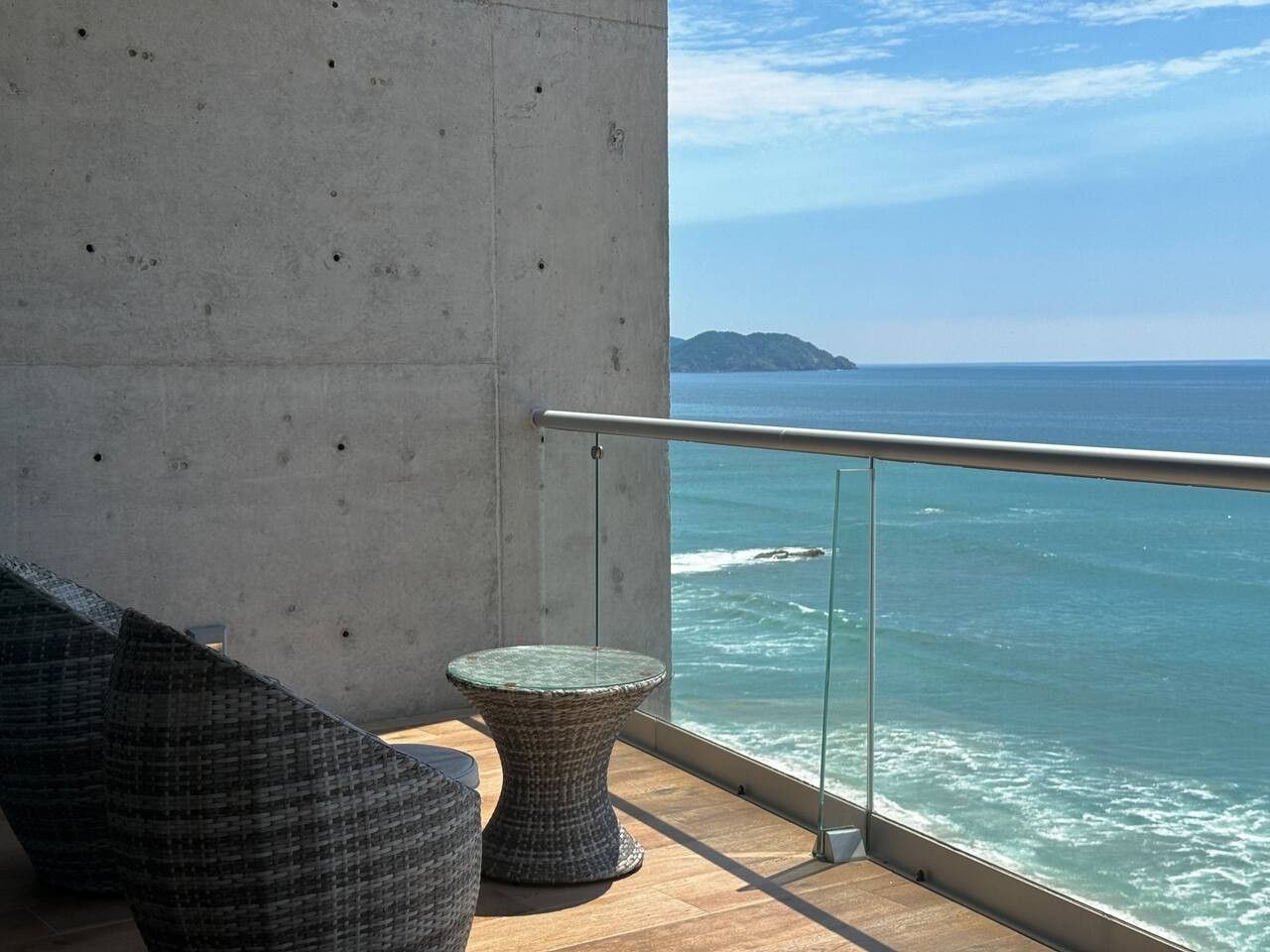 En balkon med en betonvæg, gelænder af glas og udsigt over havet.