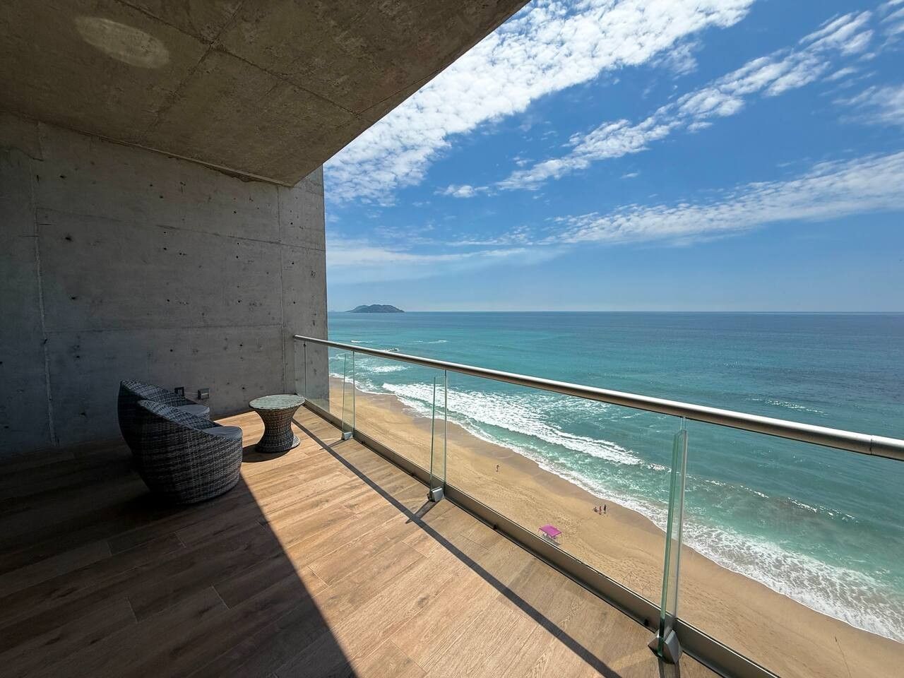 Un balcón con vista a la playa y al océano, una pared de concreto y una silla de mimbre.
