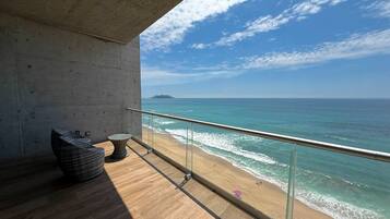 Un balcón con vista a la playa y al océano, una pared de concreto y una silla de mimbre.