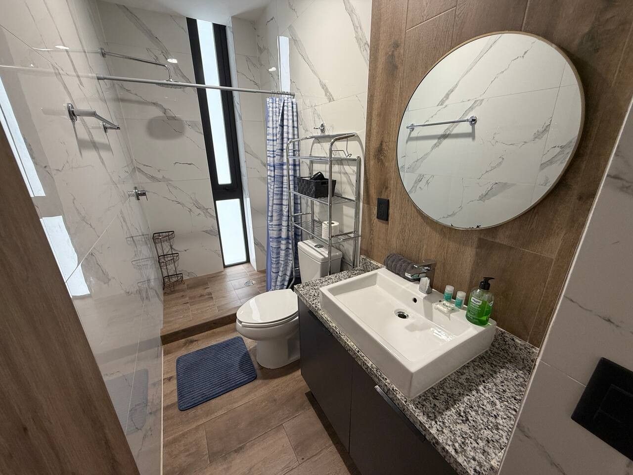 A modern bathroom with a marble floor, a round mirror, a sink with toiletries, and a shower area.