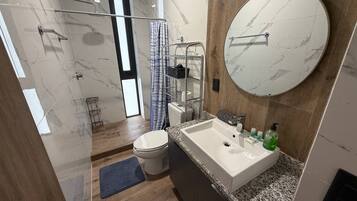 A modern bathroom with a marble floor, a round mirror, a sink with toiletries, and a shower area.