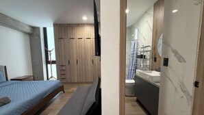 Ein Schlafzimmer mit Bett, Sitzbank, Kleiderschrank und Blick ins Badezimmer mit Waschbecken und Spiegel.
