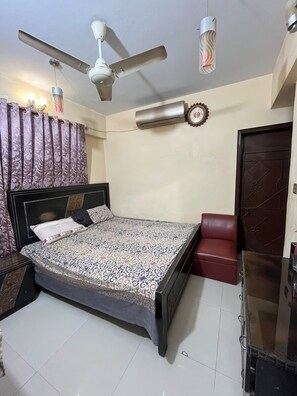 Un dormitorio con cama, ventilador de techo, aire acondicionado, una silla roja y un armario.
