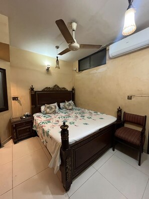Un dormitorio con cama, ventilador de techo, aire acondicionado de pared, mesita de noche, lámpara y una silla.