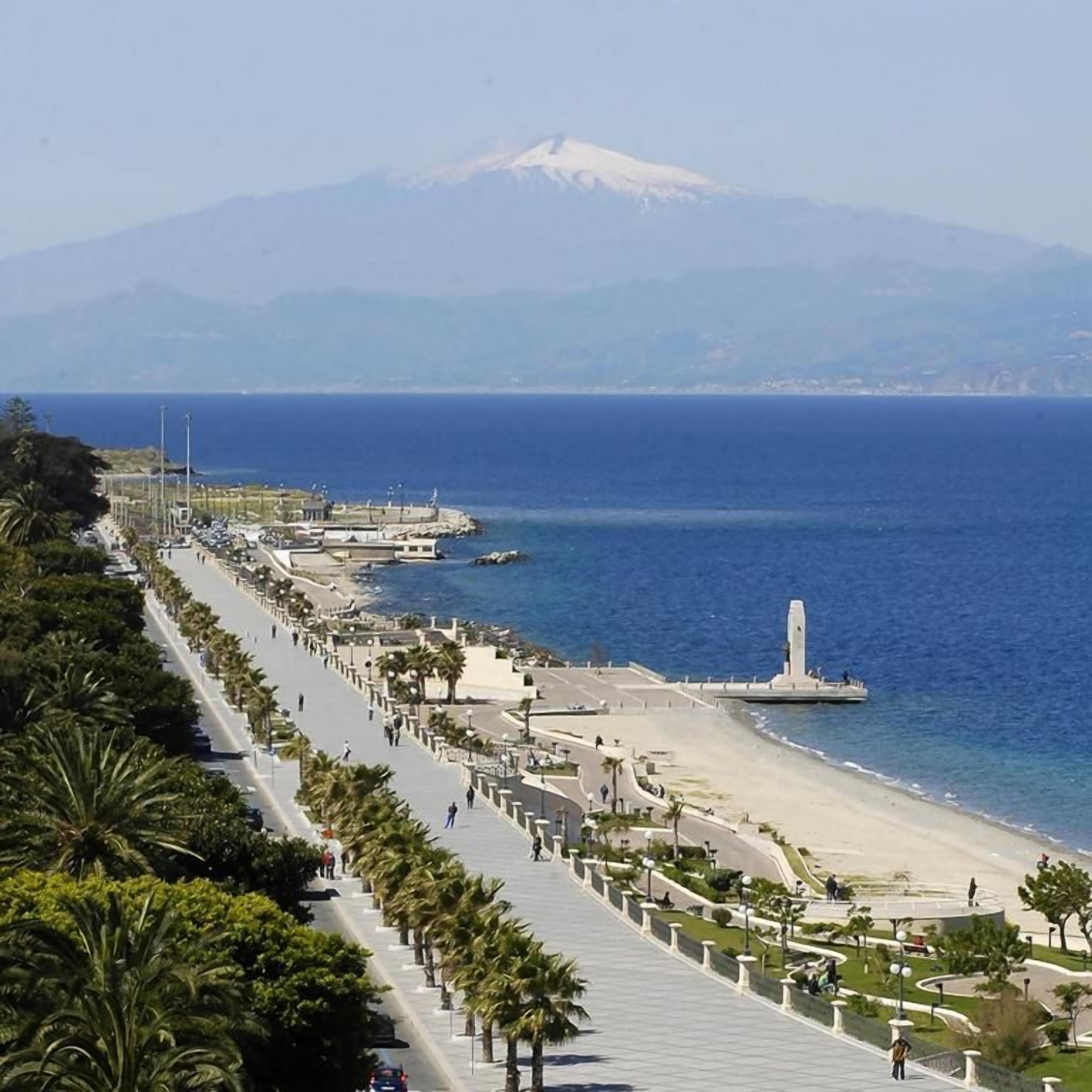 A coastal promenade with a mountain in the background, a beach, and a clear sky.