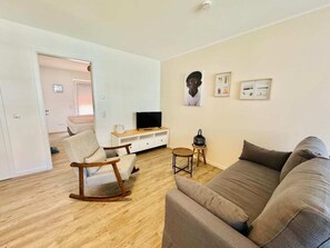 A living room with a sofa, a rocking chair, a TV on a wooden stand, and a small table with a lantern. - Beachcomber F3norderney (Norderney)