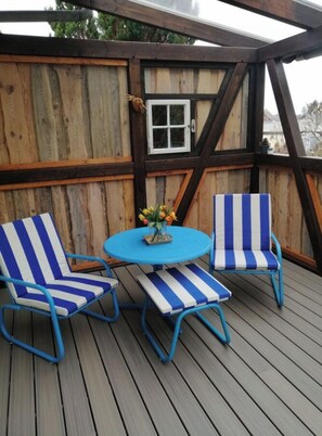 A wooden deck with a blue table and striped chairs, a small window, and a decorative item on the wall. - Holiday Apartment With 1 Bedroom (Lübbenau/Spreewald)