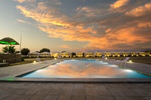 Una piscina con sillones y sombrillas, rodeada de edificios con luces.
