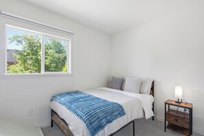 A bedroom with a bed, a bedside table with a lamp, and a window with a view of trees. - Modern 4BR Home - Parking - Close to Downtown (Seattle)