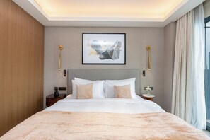 Ein Hotelzimmer mit einem großen Bett, einem gerahmten Kunstwerk darüber, Nachttischlampen, einem Nachttisch mit Telefon und einem Fenster mit Vorhängen.