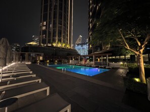 Uma área de piscina à noite, com espreguiçadeiras e guarda-sóis, cercada por edifícios com janelas iluminadas.