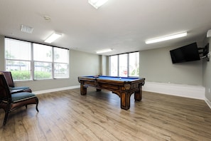 A pool table in a room with wooden flooring, large windows, and a television mounted on the wall. - Serenity at The Opus (Daytona Beach)
