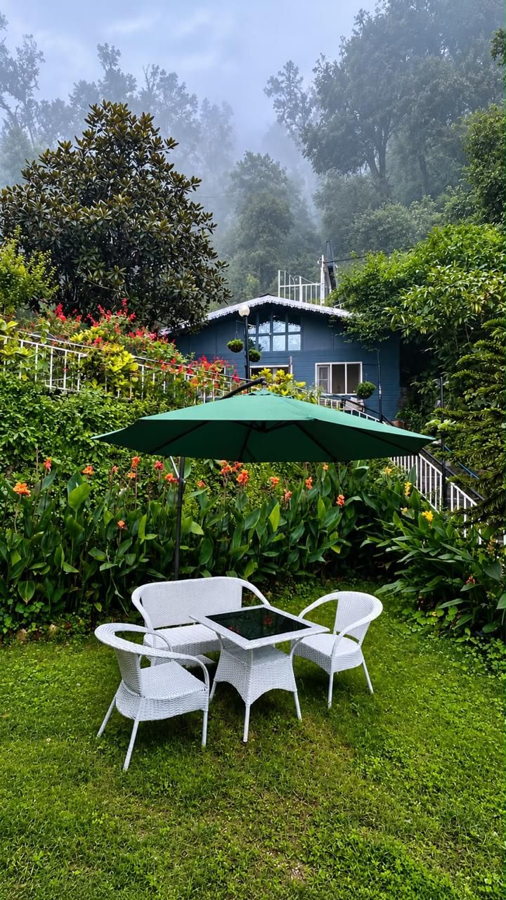 Een tuin met een witte rieten tafel en stoelen, een groene parasol en een blauw huis met een witte hekken in de achtergrond.