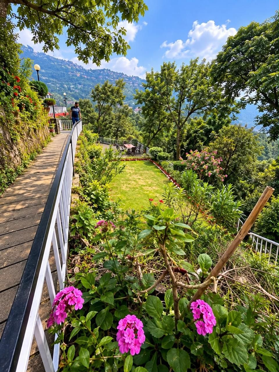 Un sendero con barandilla, rodeado de follaje y flores, que conduce a una zona de descanso con vistas a un paisaje montañoso.