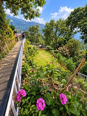 A pathway with a railing, surrounded by greenery and flowers, leading to a seating area with a view of a mountainous landscape. - The Sky Hotels (Mussoorie)