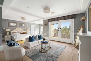 Une chambre spacieuse avec un grand lit, un coin salon comprenant un canapé et un fauteuil, une petite table ronde et un balcon aménagé avec du mobilier d’extérieur.