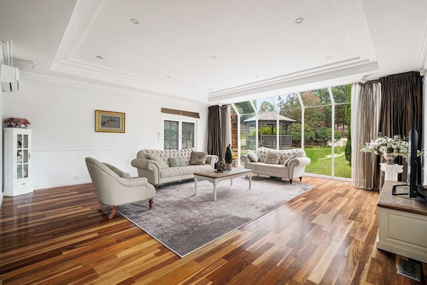 A spacious living room with a large window, a white fireplace, and a dining area with a table and chairs. - Shalom Garden Luxury Estate · Pool, Tennis Court (Melbourne)