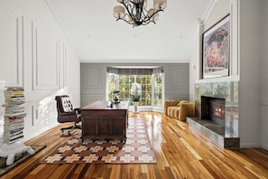 A spacious living room with hardwood flooring, a dark wooden desk, a leather chair, a fireplace, and a large window with a view of greenery.