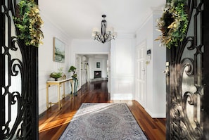 A hallway with a patterned rug, a chandelier, and a fireplace in the background.