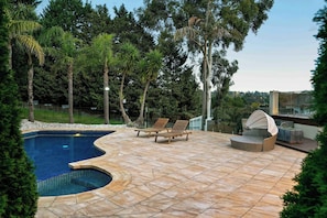A swimming pool with a curved edge, surrounded by a patio with tiled flooring and two wooden lounge chairs.