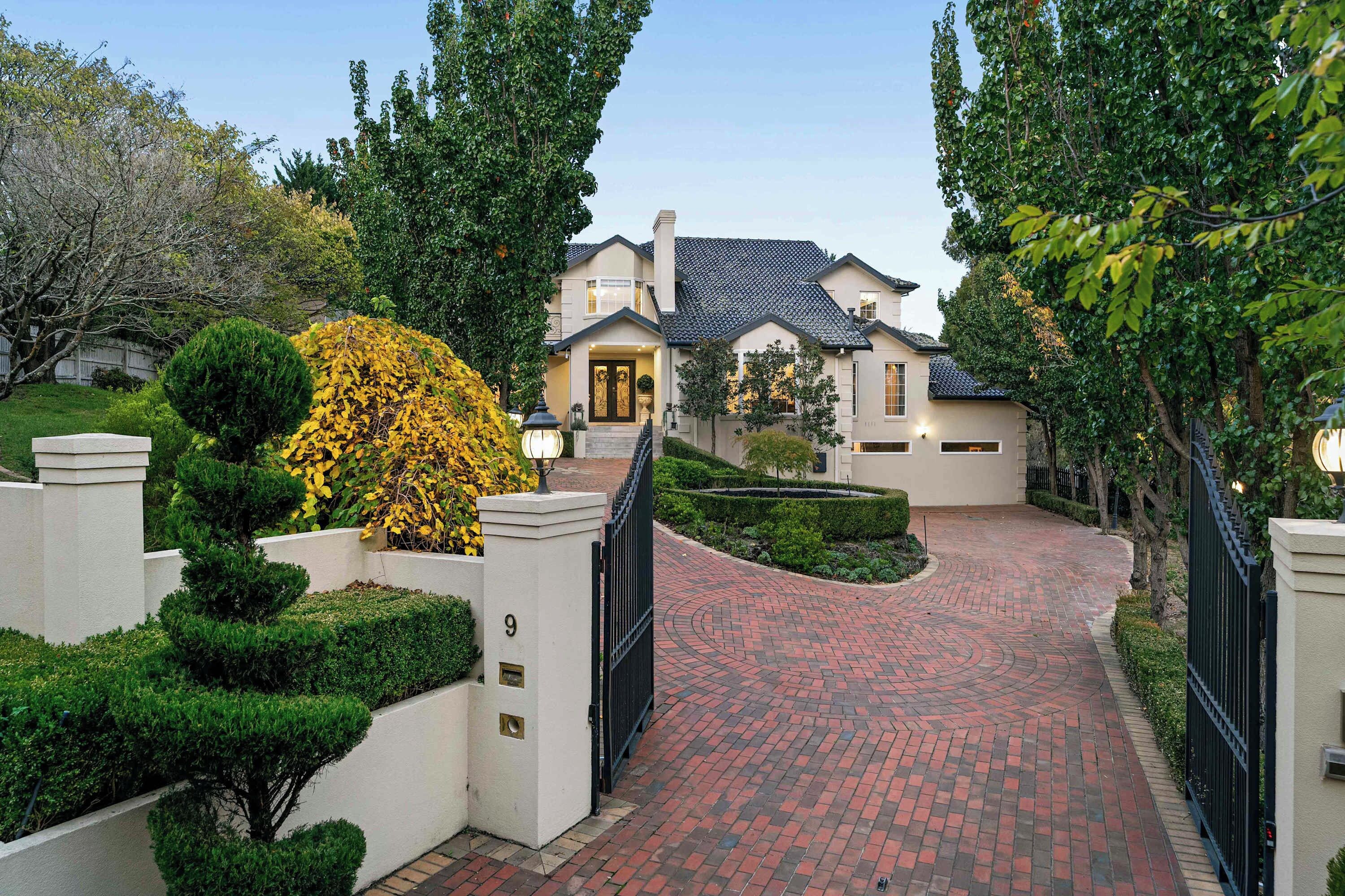 A two-story house with a brick driveway leading to a gated entrance.