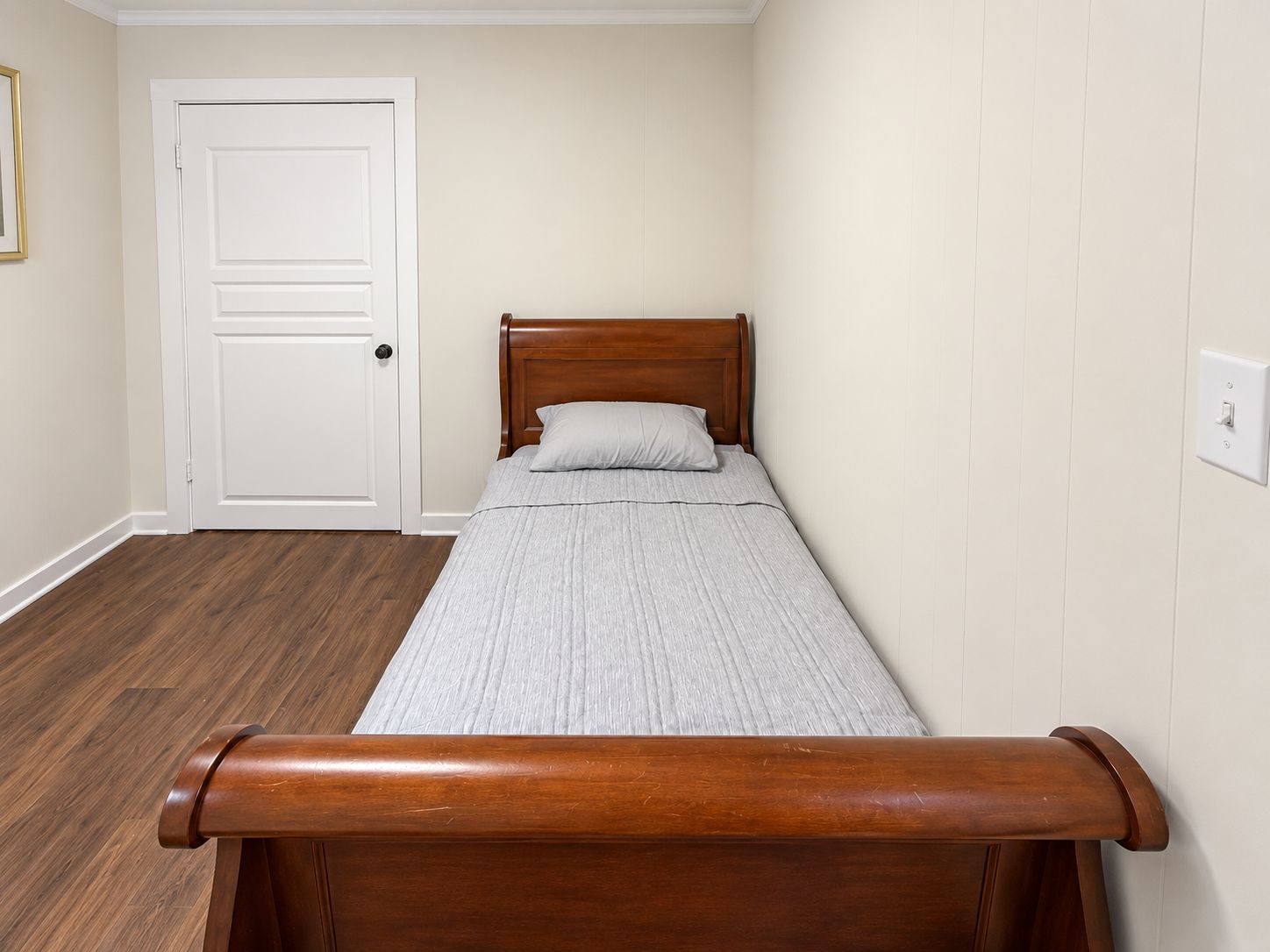Un lit simple avec une tête de lit en bois, dans une pièce dont la porte est blanche et les murs de couleur claire.