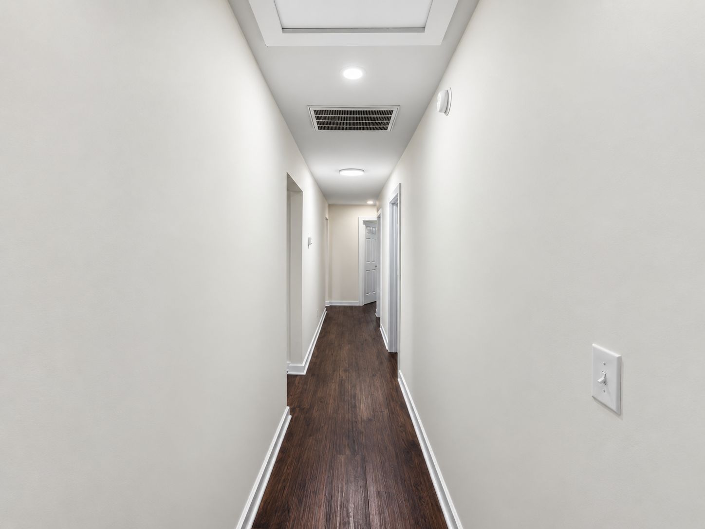 A corridor with white walls, a wooden floor, and a ceiling with recessed lighting and a vent.
