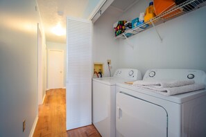A laundry room with a washing machine and dryer, a shelf with laundry supplies, and a door leading to another room. - Escape to Eden | Sunset Views & Dockage (Big Pine Key)