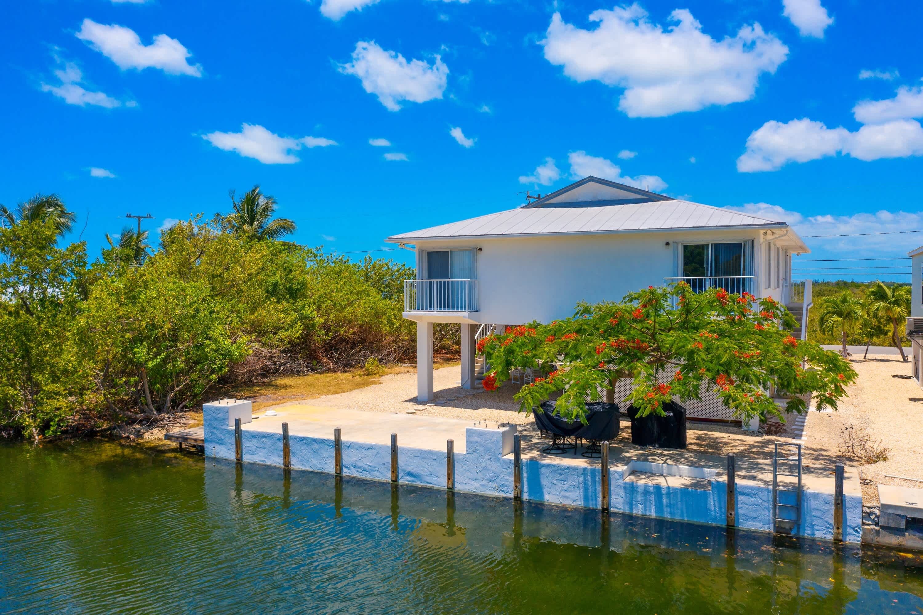 A single-story house with a balcony, situated on a waterfront with a dock extending into the water.