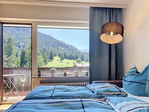 A bedroom with a bed, a window with a view of a forested mountain, and a hanging lamp. - Edelweiss by Interhome (Farchant)