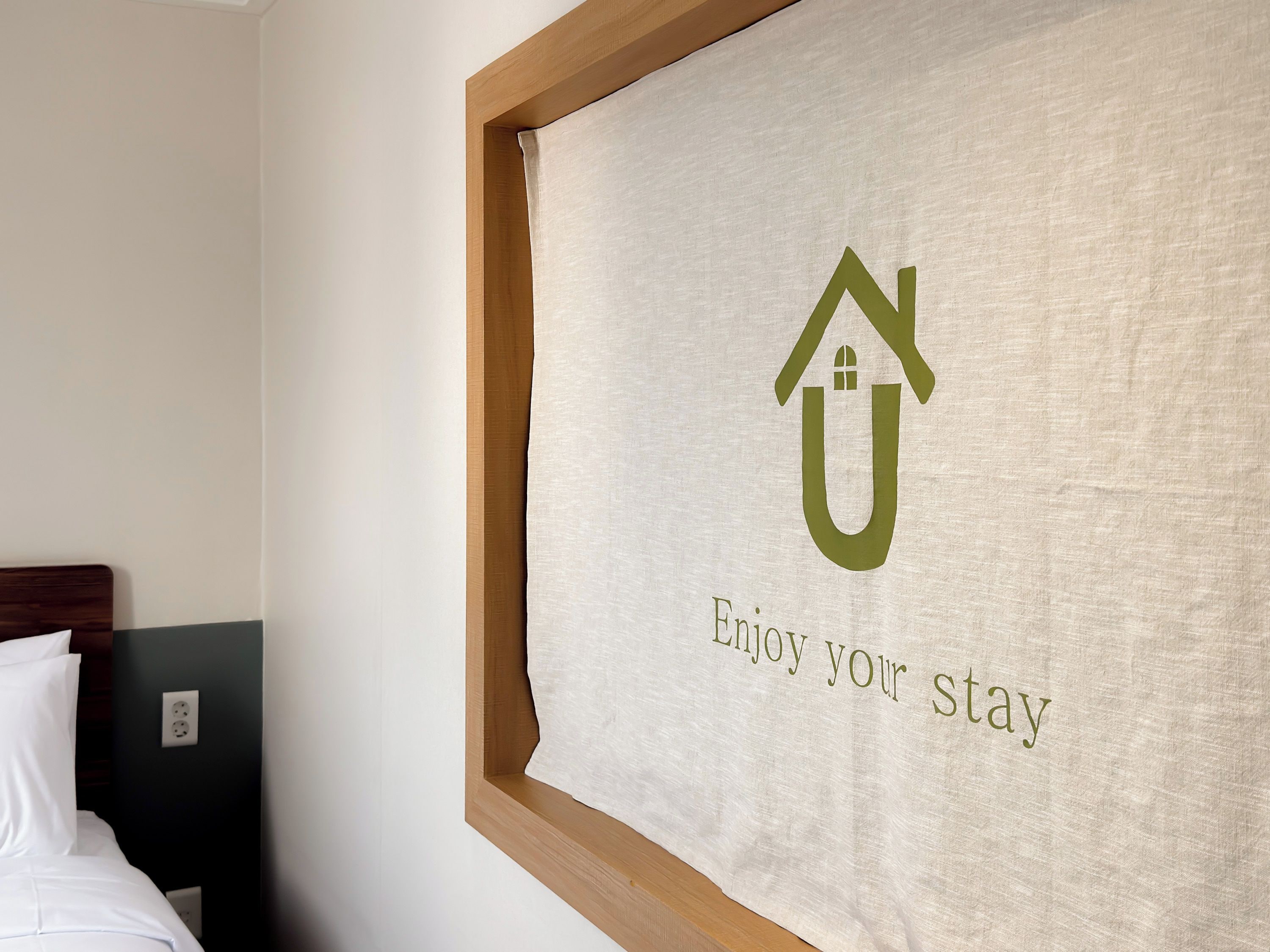 Habitación de hotel con cama, cabecera, interruptor de luz en la pared y cortina con el logotipo de una casa y el texto 'Disfruta tu estancia'.